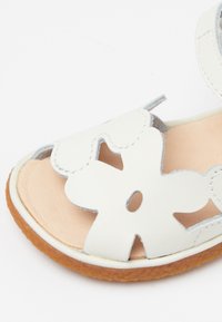 Valkoinen nahkasandaali, jossa on kukkakuvioiset leikkaukset, pehmeä beige pohjallinen, kumipohja ja harmaat ompelusaumat. Kompakti ja litteä muoto.