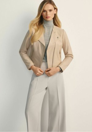 Kobieta z długimi blond włosami, ubrana w beżową skórzaną kurtkę, szary golf i jasnoszare spodnie o szerokich nogawkach, stojąca na jednolitym tle.