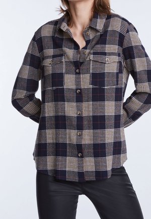Femme portant une chemise à carreaux bleu marine, beige et marron avec deux poches avant et un pantalon en cuir noir, debout les mains dans le dos.