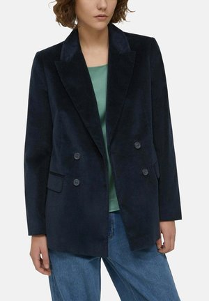 Femme portant un blazer en velours côtelé bleu marine foncé à double boutonnage sur un haut vert avec un jean bleu, debout devant un fond clair uni.