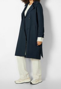 Trench coat blu navy con design doppiopetto, ampi risvolti e dettagli con bottoni. Abbinato a pantaloni chiari e scarpe da ginnastica.