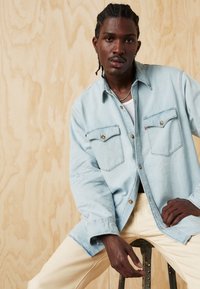 Light blue denim shirt with two front pockets and gold hardware, worn open over a white tank top, paired with cream-colored pants.