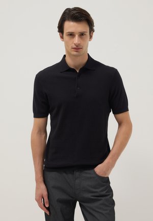 Jonge man die een zwart poloshirt met korte mouwen en een donkere broek draagt, staat met één hand in zijn broekzak tegen een effen achtergrond.