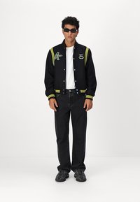 Veste de sport noire avec des accents verts et jaunes, ornée d'un patch de joueur de tennis, associée à une chemise blanche et un jean noir.