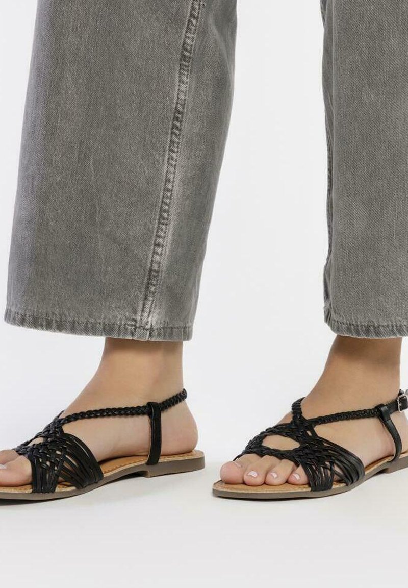 Las sandalias negras de tejido presentan un diseño de punta abierta con tiras trenzadas y un cinturón en el tobillo. La suela es de color marrón claro, que complementa la parte superior negra.
