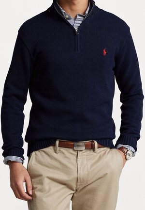 Mann trägt dunkelblauen Pullover mit Viertelreißverschluss über einem gestreiften Hemd mit Kragen, beige Hose mit braunem Gürtel und silberner Armbanduhr.