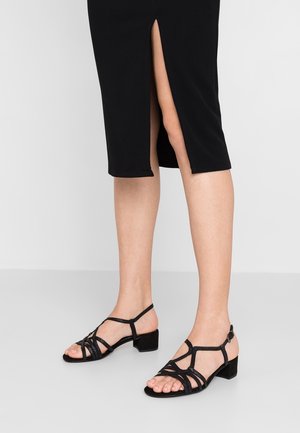 Jambes portant des sandales noires à brides et à talons bas, et une jupe noire fendue à l'arrière, debout sur un sol gris clair.