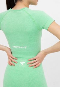 Žena ve světle zeleném sportovním oblečení s logem "SQUATPROOF" na zádech, s rukama opřenýma o dolní část zad a pas proti bílé zdi.