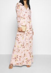 Abito lungo floreale in rosa chiaro con maniche lunghe, scollatura a V profonda e vita con cintura. Indossato con una pochette dorata testurizzata e sandali con cinturini.