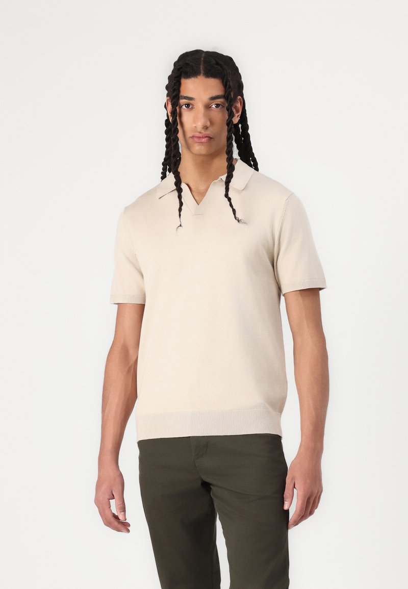 Beige poloko s krátkým rukávem, V-výstřihem a žebrovaným lemem, ve spojení s tmavě zelenými kalhotami. Hladká textura, přiléhavý střih.