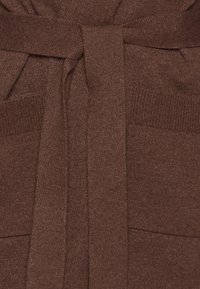 Brązowy sweter z dzianiny z paskiem do zawiązania, łatami na kieszeniach i ściągaczami na rękawach. Tkanina ma gładką, miękką teksturę.