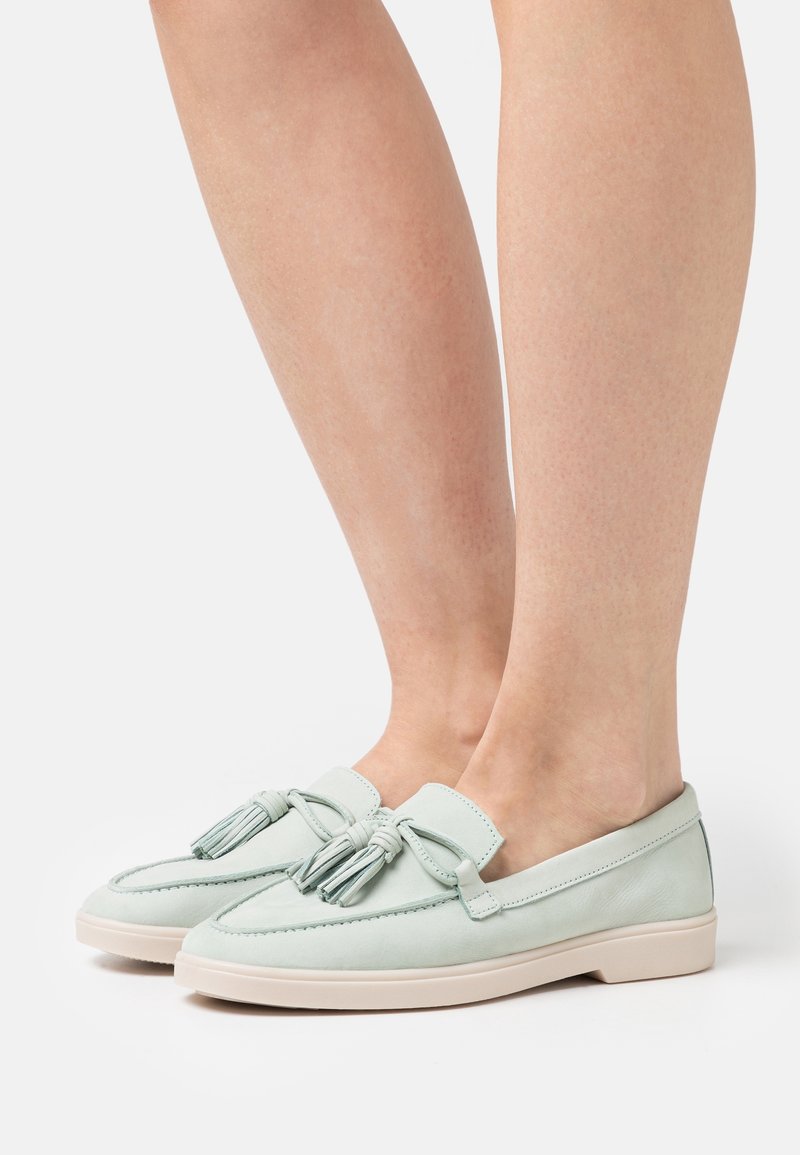 Mocassins en cuir vert clair avec pompons, bout arrondi, surpiqûres contrastées et semelle en caoutchouc beige. Portés sur jambes nues.