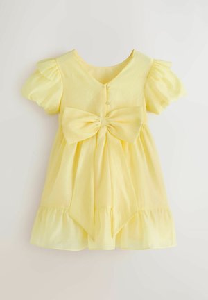 Dzeltena kleita ar īsām volānu piedurknēm, V veida izgriezumu un lielu priekšējo loku. Svārkiem ir savilkts apakšmala, lai piešķirtu apjomīgumu.