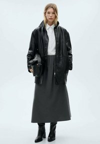 Schwarze Lederjacke über einem weißen Hemd, kombiniert mit einem grauen langen Rock und knielangen schwarzen Stiefeln, was einen monochromatischen Look schafft.