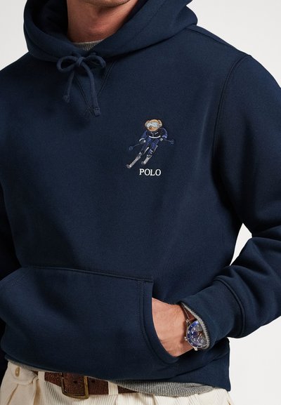 Marineblå hettegenser med brodert skiløper og «POLO»-tekst på brystet, frontlomme, brukt over en grå genser, med blå klokke synlig på håndleddet.