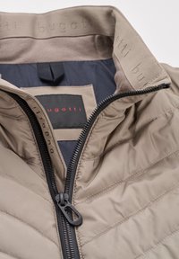 Beige Daunenjacke mit gestepptem Muster, schwarzem Reißverschluss und Bugatti-Logo. Das Innenfutter besteht aus dunklem Stoff und hat eine gebrandete Kragenverzierung.