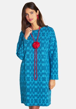 Türkises, strukturiertes Kleid mit langen Ärmeln und geometrischem Muster. Eine rote Blütenkette und passende Ohrringe runden den Look ab.