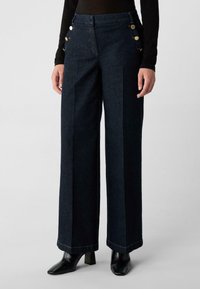 Kobieta ubrana w ciemne, szerokie dżinsy z złotymi guzikami po bokach, czarne botki na obcasie i czarną bluzkę z długim rękawem na jednolitym tle.