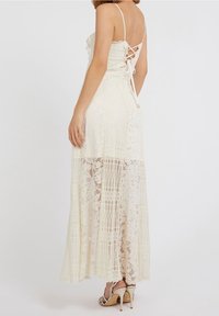 Robe longue en dentelle crème avec un design à étages et des bretelles fines. Présente un dos lacé et une jupe fluide. Portée avec des talons à lanières.