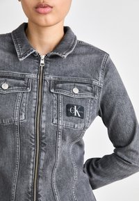 Grå denimjakke med kragehals, glidelås, brystlommer og et Calvin Klein Jeans-logo-patch på venstre side.