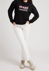Sudadera negra con el logo "GUESS Originals" en rojo y blanco, combinada con pantalones blancos y botas beige con cordones. Tejido suave y corte relajado.