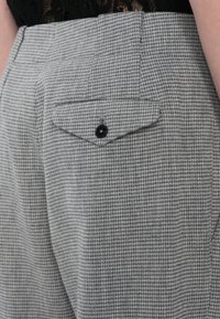 Pantalon gris à motif pied-de-poule avec un rabat de poche arrière sécurisé par un bouton noir, porté avec un vêtement noir rentré.