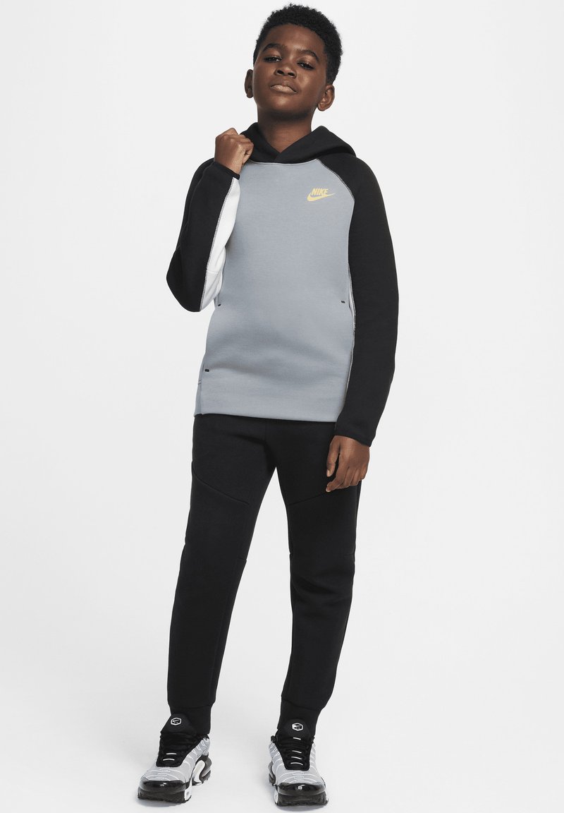 Sudadera de Nike en gris y negro con capucha, que presenta un logotipo amarillo. Combinada con pantalones jogger negros y zapatillas negras/plateadas. Textura suave.