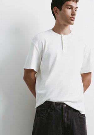 Ung mand iført en hvid henley med korte ærmer og sorte jeans, stående med hænderne bag ryggen, kiggende til siden mod en ensfarvet baggrund.