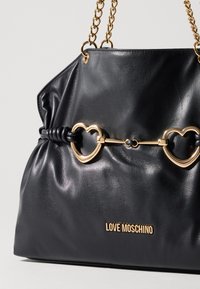 Black faux leather handbag with a gathered design, gold heart-shaped hardware, and chain straps. Features a logo at the bottom.