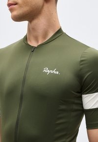 Άνδρας που φοράει ποδηλατική φανέλα Rapha σε ελαιζένιο πράσινο με φερμουάρ μπροστά και λευκή ταινία στο κοντό μανίκι, με απλό φόντο.
