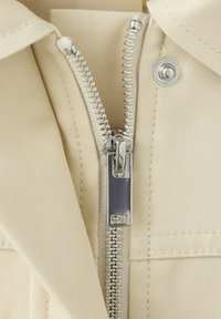 Zbliżenie na srebrny metalowy zamek błyskawiczny częściowo zapięty na beżowej tkaninie z widocznym przeszyciem i okrągłym metalowym zatrzaskiem.