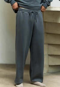 Los pantalones de chándal grises cuentan con una cintura elástica, cordón y bolsillos laterales. La tela parece suave con una textura lisa y un diseño de pierna ancha.