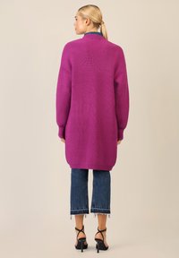 Długi różowy sweter z dzianiny o ryflowanej fakturze, otwarty przód, obniżone ramiona i długie rękawy; w połączeniu z krótkimi dżinsami i czarnymi szpilkami.