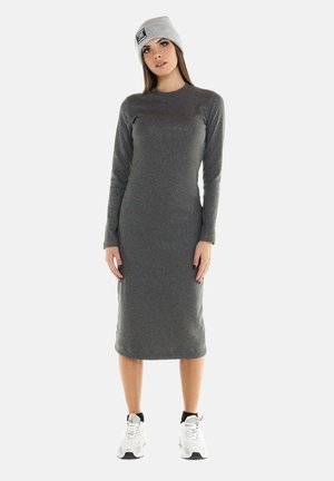 Jeune femme portant une robe grise à manches longues, longueur genoux, un bonnet gris clair et des baskets blanches avec des chaussettes noires, debout sur fond blanc.