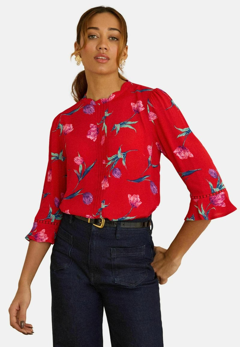 Naine, kellel on punane lilleline pluus lõhestatud kaelusega ja kolmveerand pikkused satsilised varrukad, kombineeritud tumedate kõrge vöökohaga teksade ja musta vööga.