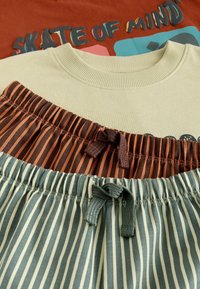 Chemise beige avec le texte "SKATE OF MIND" partiellement visible, associée à un pantalon à taille élastique rayé marron et bleu avec des cordons.