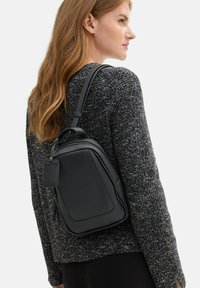 Sac à dos en cuir noir de forme rectangulaire, avec une texture lisse et des bretelles ajustables ; présente une petite étiquette sur le côté.
