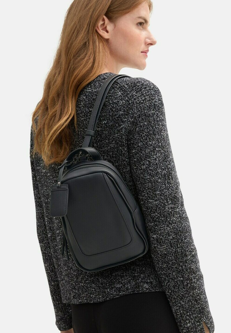 Sac à dos en cuir noir de forme rectangulaire, avec une texture lisse et des bretelles ajustables ; présente une petite étiquette sur le côté.