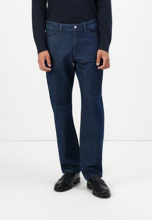 Man wearing dark blue straight-leg jeans, black loafers, and a long-sleeve navy sweater against a plain white background.