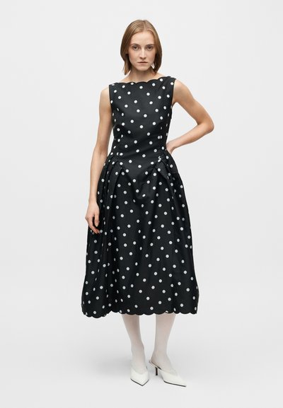 Svart klänning med vita prickar med figursydd överdel och veckad kjol. Har en vågig halsringning och texturerat tyg. Bärs med vita klackar.