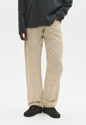 Osoba nosząca beżowe szerokie spodnie jeansowe, czarną koszulę z długim rękawem i czarne trampki, stojąca z jedną ręką w kieszeni.