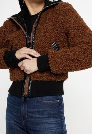 Persona vistiendo una chaqueta marrón con cremallera y textura, con puños y cinturilla negros, combinada con vaqueros azules, con las manos entrelazadas a la altura de la cintura.