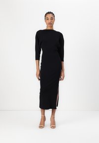 Robe en tricot côtelé noir avec un décolleté bateau et des manches trois-quarts, présentant une fente sur le côté et une silhouette ajustée, associée à des talons nus à lanières.