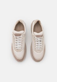 Ett par beige och vita mockasneakers med vita snören, sedd uppifrån på en vit bakgrund.