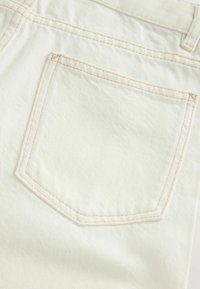 Le tissu en denim blanc présente une poche arrière rectangulaire avec des coutures brun clair et une surface légèrement texturée. Design simple, sans embellissements.