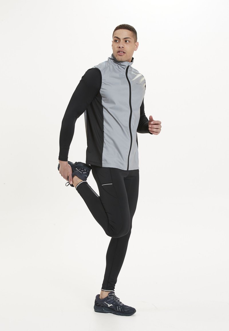 Athletisches Trikot aus grauem und schwarzem Stoff, mit einem Reißverschluss vorne, kombiniert mit schwarzen Leggings und dunklen Laufschuhen. Flaches, elegantes Design.