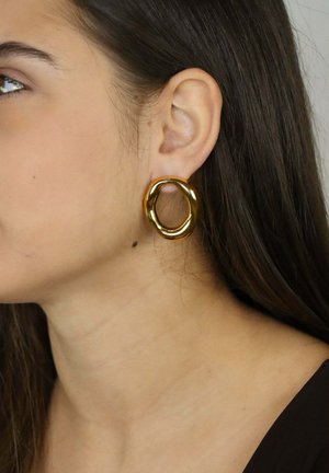 Goldener, skulpturaler Creolen-Ohrring in ovaler Form mit glatter, glänzender Oberfläche, präsentiert neben dem Ohr einer Person.