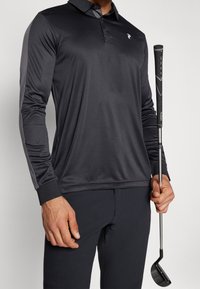 Polo golf a maniche lunghe nera in un tessuto morbido e leggero, con un colletto e dettagli grigi sulle maniche. Una mazza da golf è tenuta nella mano destra.