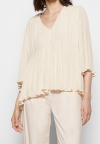Cremefarbene, plissierte Bluse mit V-Ausschnitt, fließender Silhouette und gerafftem Saum. Leichter Stoff, lange Ärmel und weiche Textur, kombiniert mit Satin-Hosen.