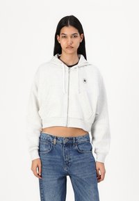 Grey cropped hoodie with a zipper and drawstrings, featuring a small logo on the chest and two front pockets. Worn with blue denim jeans.